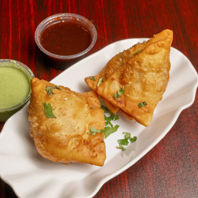 Two golden-fried samosas garnished with cilantro, served with green and red chutneys on a white plate.