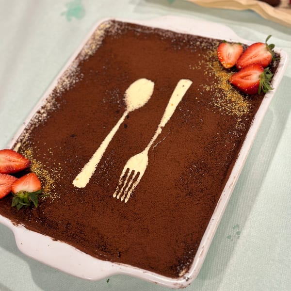 Tiramisu dessert with cocoa powder, spoon and fork design, and fresh strawberries in a white dish.