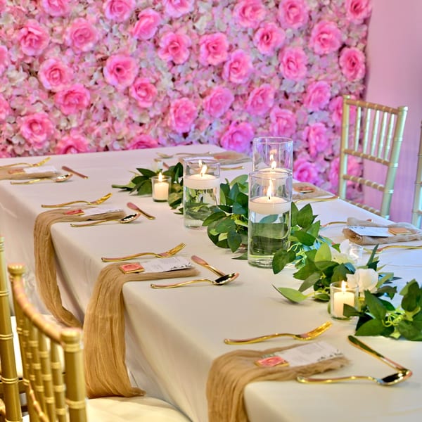 Elegant table setting with gold cutlery, candles, and greenery against a pink rose wall.