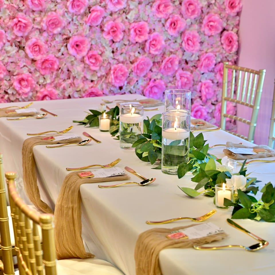 Elegant table setting with gold cutlery, candles, and greenery against a pink rose wall.