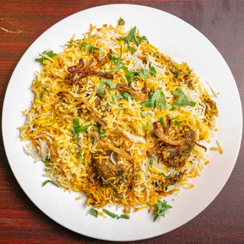 A white plate filled with Indian biryani, featuring yellow and white rice, meat, fried onions, and fresh herbs.